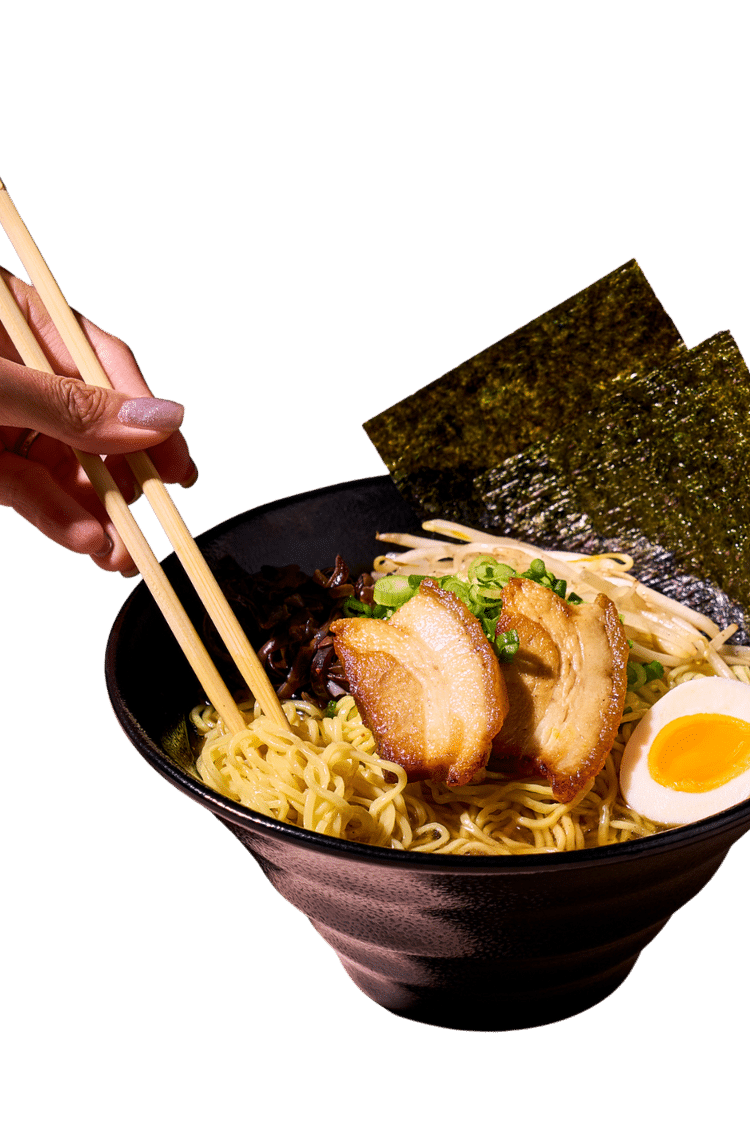 A hand with painted nails uses chopsticks to pick up noodles from a black bowl of ramen.